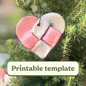Puede incluir: Un adorno en forma de corazón con un diseño tejido, con secciones alternas de rojo y gris. El adorno cuelga de una cuerda negra y se coloca sobre un fondo de agujas de pino verdes. El texto "Printable template" se muestra en una pancarta verde.