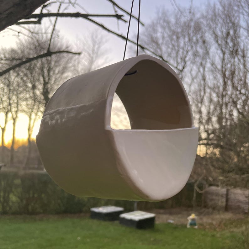 May include: A light pink ceramic hanging planter with a rounded shape and a wide opening. The planter is suspended by a black cord.