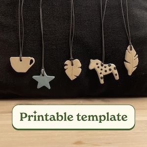 Op de afbeelding: Vijf keramische hangers aan zwarte koorden, waaronder een kopje, een ster, een blad, een paard en een veer. De hangers zijn beige en blauw met zwarte accenten. De tekst "Printbare sjabloon" staat onderaan de afbeelding.