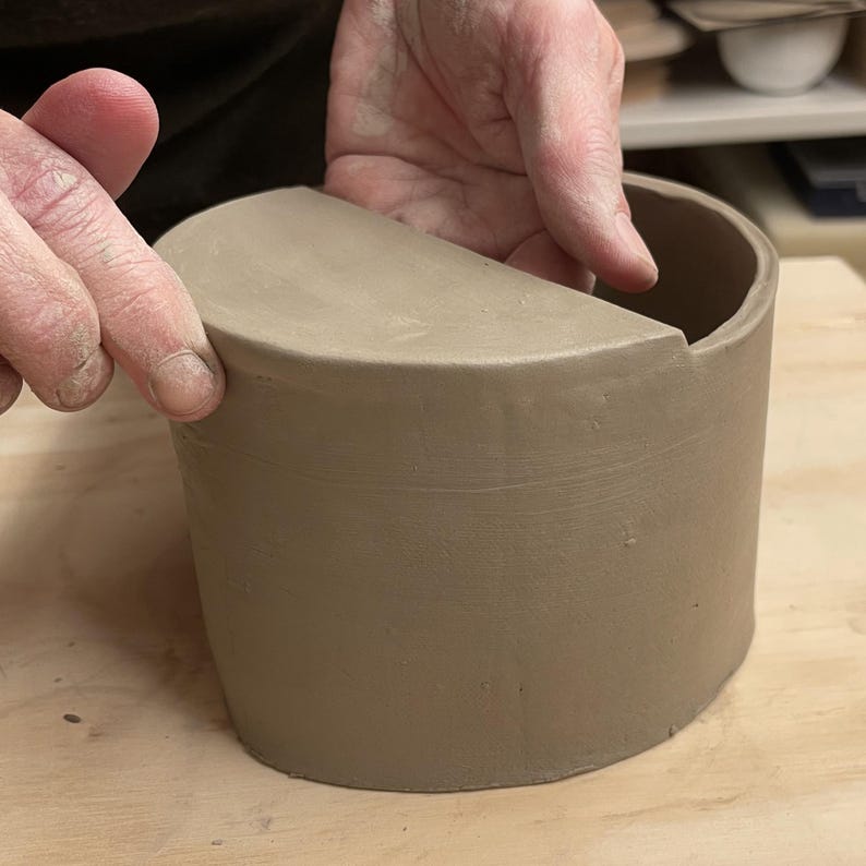 May include: A brown ceramic pot with a half-circle cut out on the top. The pot is being held by a hand.
