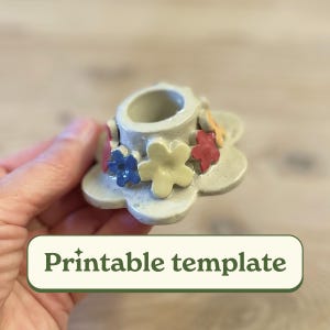 Op de afbeelding: Een witte keramische kaarsenhouder met een bloemenontwerp. De bloemen zijn geel, rood, blauw en oranje. De houder heeft een ronde opening in het midden voor een kaars.