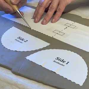 May include: A person uses a tool to cut out a paper pattern for a bird feeder. The pattern is labeled "Side 1 Birdfeeder" and "Side 2 Birdfeeder".