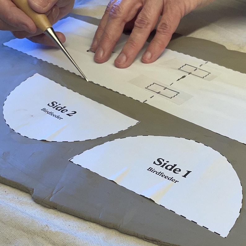 May include: A person uses a tool to cut out a paper pattern for a bird feeder. The pattern is labeled "Side 1 Birdfeeder" and "Side 2 Birdfeeder".