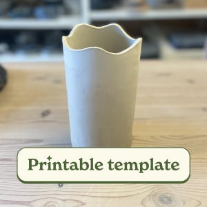 Peut inclure: Un vase en céramique gris clair avec un bord supérieur ondulé. Le vase est cylindrique et repose sur une surface en bois clair. Le texte "Printable template" est dans un rectangle vert en dessous.