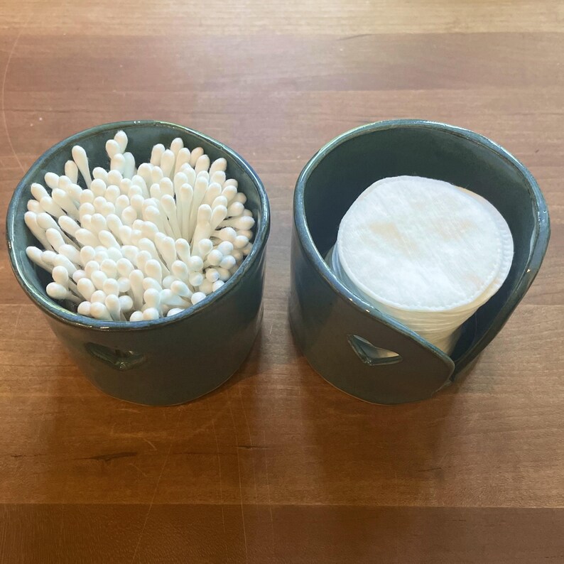May include: Two teal ceramic containers, one filled with white cotton swabs and the other with white cotton pads. Both containers have a heart-shaped cutout on the side. The containers sit on a wooden surface.