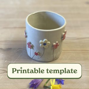 Può includere: Una tazza in ceramica bianca con un motivo floreale. La tazza ha fiori rosa, blu e gialli con steli verdi.