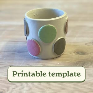 Puede incluir: Maceta de cerámica blanca con un patrón de lunares. Los lunares son en tonos de rosa, verde y marrón.