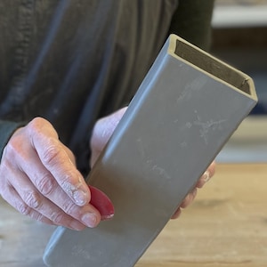 May include: A gray ceramic vase with a rectangular shape. The vase is being held by a person's hand.
