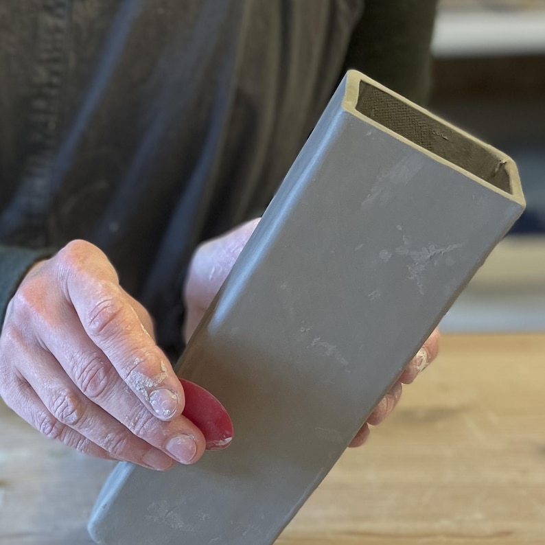 May include: A gray ceramic vase with a rectangular shape. The vase is being held by a person's hand.