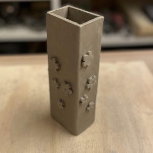 May include: A brown ceramic vase with a rectangular shape and a textured surface. The vase has a series of small flower-shaped impressions on the front.
