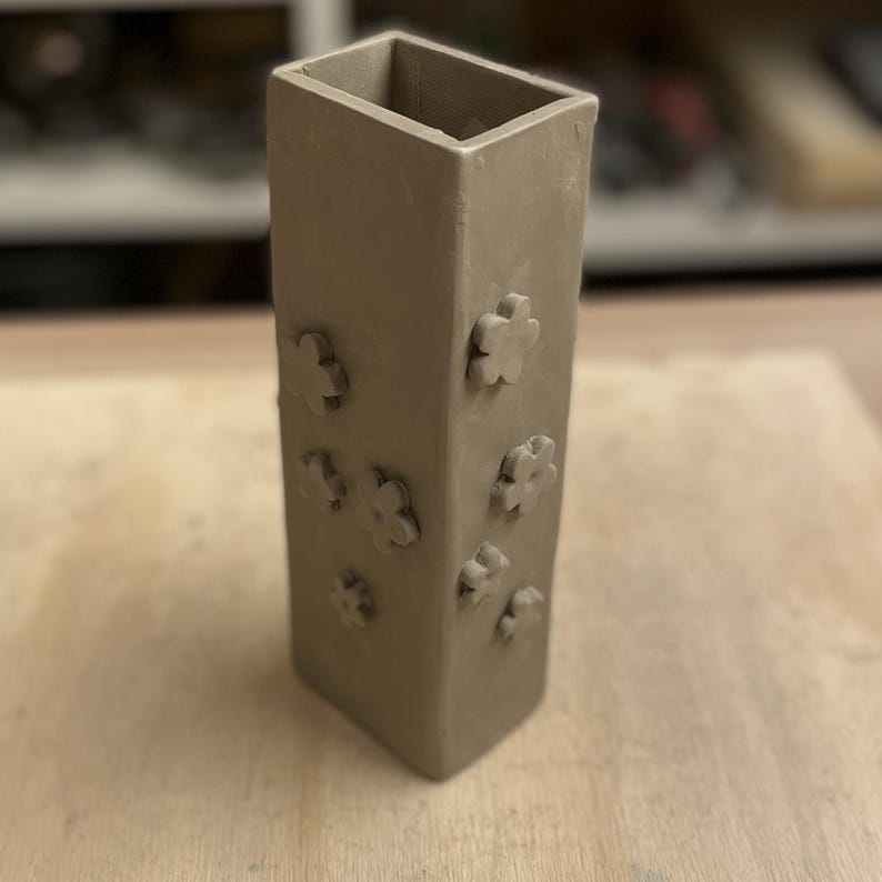 May include: A brown ceramic vase with a rectangular shape and a textured surface. The vase has a series of small flower-shaped impressions on the front.