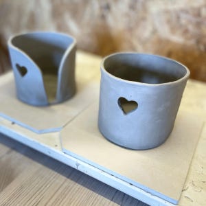 May include: Two handmade, gray ceramic containers. One is a small, open-sided holder with a heart-shaped cutout. The other is a small, round pot with a heart-shaped cutout. Both are on a light-colored surface.