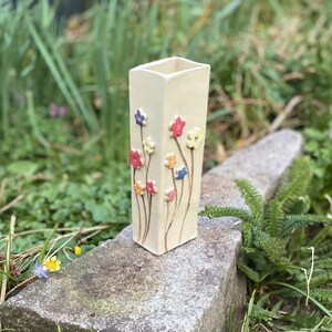 May include: A white ceramic vase with a square shape and a floral design. The vase is decorated with small, colorful flowers in shades of red, yellow, blue, and pink.