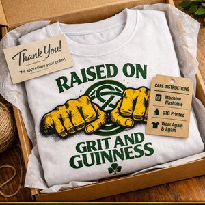 May include: A white t-shirt with the text "RAISED ON GRIT AND GUINNESS" in green, featuring a graphic of two yellow fists. The shirt is inside a cardboard box with a "Thank You" card and a care instructions tag.