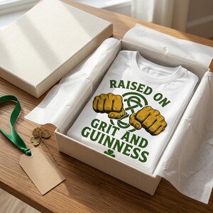 May include: White t-shirt with the text "RAISED ON GRIT AND GUINNESS" in green, featuring two fists and a Celtic knot design. The shirt is presented in a gift box with tissue paper, a green ribbon, a tag, and a four-leaf clover.