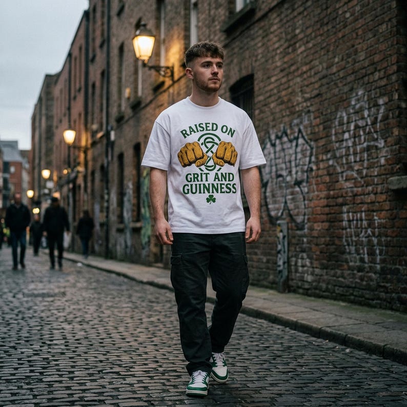 May include: A white t-shirt with the text "RAISED ON GRIT AND GUINNESS" in green and gold, featuring a shamrock graphic. The person is wearing black cargo pants and white sneakers with green accents. The background is a brick-lined street.