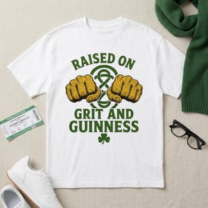 May include: White t-shirt with the text "RAISED ON GRIT AND GUINNESS" in green. The design includes two clenched fists and a Celtic knot. A shamrock is at the bottom of the design.