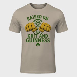 May include: A beige t-shirt with the text "RAISED ON GRIT AND GUINNESS" in green, featuring two fists and a shamrock. The fists are yellow and the text is in a bold, stylized font. The shirt is a crew neck.