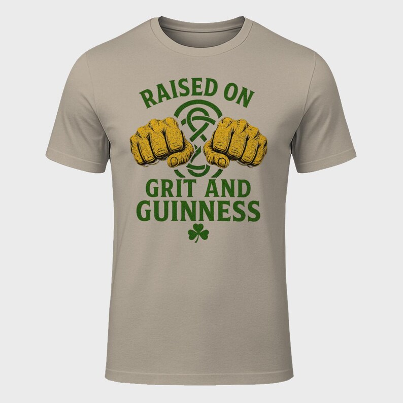 May include: A beige t-shirt with the text "RAISED ON GRIT AND GUINNESS" in green, featuring two fists and a shamrock. The fists are yellow and the text is in a bold, stylized font. The shirt is a crew neck.