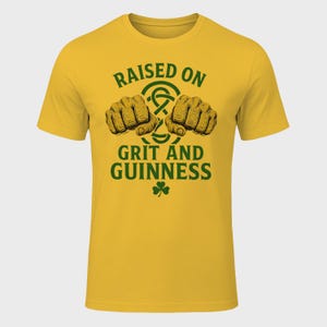 May include: Mustard yellow t-shirt with the text "RAISED ON GRIT AND GUINNESS" in green. The design includes two clenched fists, a Celtic knot, and a shamrock. The shirt is a short-sleeved crew neck.