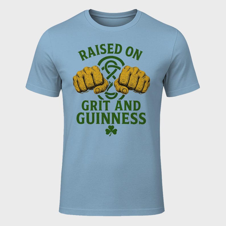 May include: Light blue t-shirt with the text "RAISED ON GRIT AND GUINNESS" in green. The design includes two fists, a Celtic knot, and a shamrock. The shirt is short-sleeved and made of a soft material.