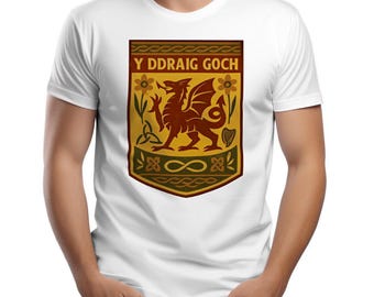May include: A white short-sleeved t-shirt featuring a central shield graphic. The shield displays a red dragon, yellow flowers, and the text Y DDRAIG GOCH in a dark red banner. Below the dragon, a green section contains an infinity symbol.
