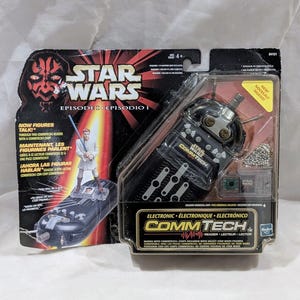 Puede incluir: Lector CommTech Star Wars Episodio I, un dispositivo electrónico negro con una placa frontal y botones plateados. El empaque presenta un personaje de Darth Maul rojo y negro y un personaje Jedi blanco. El texto en el empaque dice "Star Wars Episodio I Episodio I" y "CommTech Reader - Lecteur - Lector".