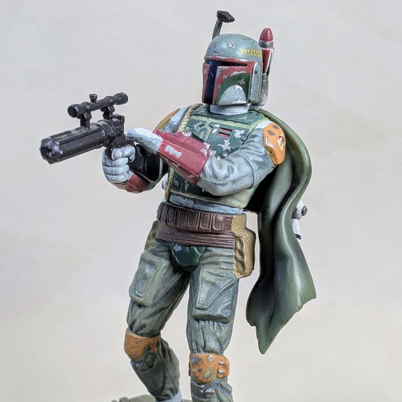 Pu&ograve; includere: Una dettagliata action figure di Boba Fett, personaggio di Star Wars. La figura &egrave; in posa di combattimento, con un fucile blaster. Presenta una combinazione di colori verde, rosso e grigio, con un mantello verde. La figura &egrave; molto dettagliata.