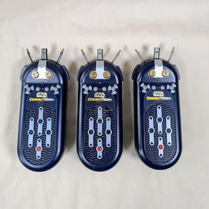 Puede incluir: Tres walkie-talkies azules Star Wars CommTech con detalles amarillos y plateados. Cada walkie-talkie tiene un panel de botones negro y plateado con un botón rojo en el centro.