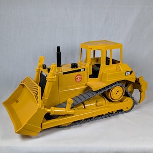 May include: A yellow Caterpillar bulldozer toy with black tracks and a front blade. The toy has a cab with windows and the CAT logo is visible on the side. The toy is made of plastic.