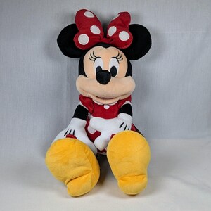 May include: A plush Minnie Mouse doll, featuring a red dress with white polka dots, a matching bow, and yellow shoes. The doll has black ears, a black nose, and white gloves. The doll is sitting down.