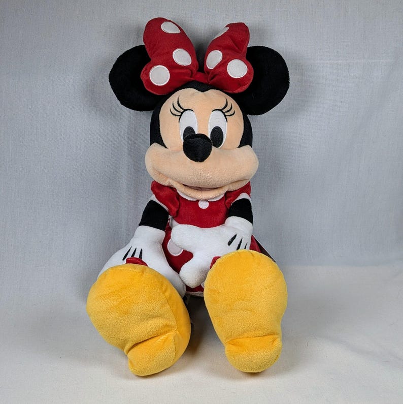 May include: A plush Minnie Mouse doll, featuring a red dress with white polka dots, a matching bow, and yellow shoes. The doll has black ears, a black nose, and white gloves. The doll is sitting down.