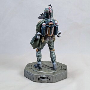 Pu&ograve; includere: Statuetta dettagliata di Boba Fett, vista da dietro. La figura indossa un'armatura verde, grigia e marrone, con un mantello verde. Si trova su una base esagonale grigia. La figura ha un jetpack e accessori per armi.