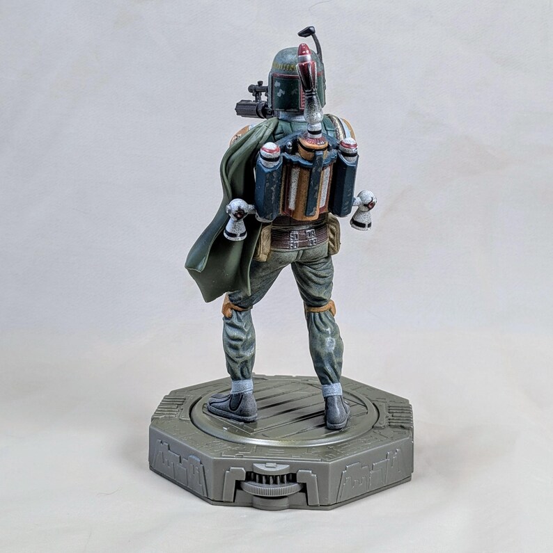 Pu&ograve; includere: Statuetta dettagliata di Boba Fett, vista da dietro. La figura indossa un'armatura verde, grigia e marrone, con un mantello verde. Si trova su una base esagonale grigia. La figura ha un jetpack e accessori per armi.