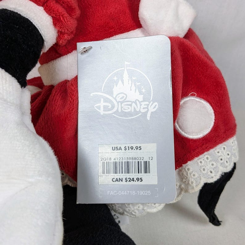 May include: A plush toy dressed in a red and white Santa Claus outfit, with a Disney tag attached. The tag features the Disney logo and product information.