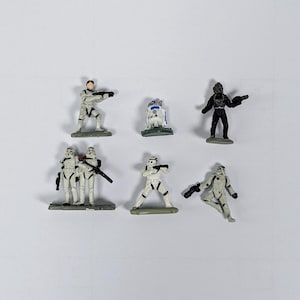 May include: A set of nine Star Wars action figures, including a Stormtrooper, a Rebel pilot, and a droid. The figures are all in white and grey, and they are standing on a white background.