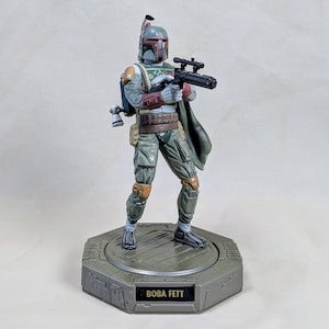 Pu&ograve; includere: Una statuetta di Boba Fett su una base grigia. La statuetta indossa un'armatura verde e grigia e tiene in mano un fucile blaster nero. Il casco &egrave; verde e rosso. La base ha il nome "BOBA FETT" in testo bianco.