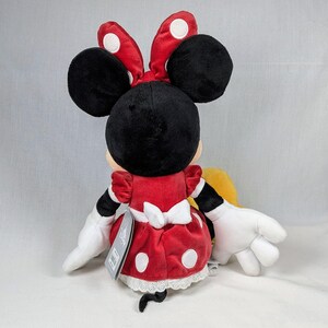 May include: A plush Minnie Mouse doll, viewed from the back, features a red dress with white polka dots and a white bow. The doll has large black ears, a red bow with white polka dots, and white gloves. A tag is attached.