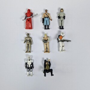 May include: A collection of nine Star Wars action figures, including a red-robed figure with a staff, a white-armored figure with a blaster, a gold-colored figure, and a black-clad figure.