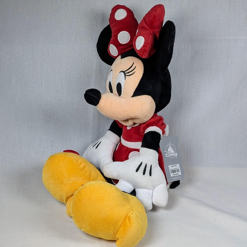 May include: A plush Minnie Mouse doll, featuring a red dress with white polka dots, a matching bow, and yellow shoes. The doll has black ears, a black nose, and white gloves. A Disney tag is attached.