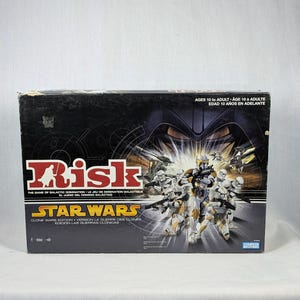 May include: A Star Wars Risk board game box. The box is black with the word "Risk" in red and white. The Star Wars logo is below the word "Risk". The box features images of clone troopers and the text "Clone Wars Edition".