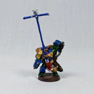 May include: A miniature figurine of a space marine, painted in blue, green, and gold armor, holding a weapon and a blue staff with a cross-like structure at the top. The figurine stands on a brown base.