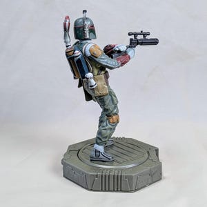 Pu&ograve; includere: Una statuetta dettagliata di Boba Fett, un personaggio di Star Wars, in una posa dinamica. La figura &egrave; vestita con un'armatura verde, grigia e rossa, che tiene in mano un fucile blaster. Si trova su una base esagonale grigia.