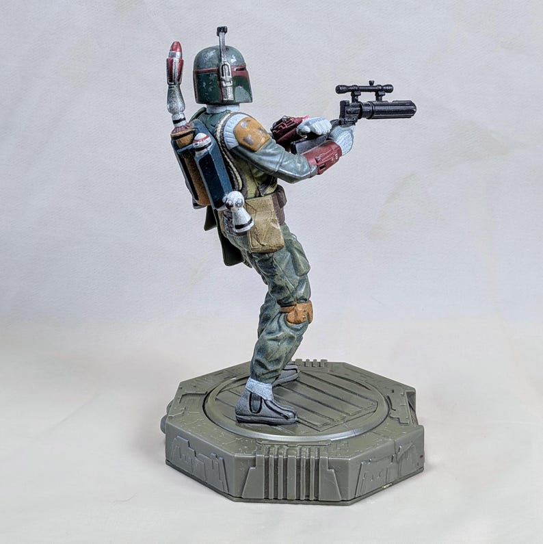 Pu&ograve; includere: Una statuetta dettagliata di Boba Fett, un personaggio di Star Wars, in una posa dinamica. La figura &egrave; vestita con un'armatura verde, grigia e rossa, che tiene in mano un fucile blaster. Si trova su una base esagonale grigia.