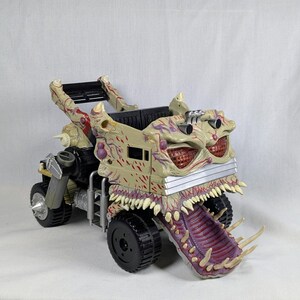 Op de afbeelding: Een speelgoed monstertruck in beige en rood, met een groot, open mondontwerp en scherpe tanden. De truck heeft zwarte wielen en een gedetailleerd, grotesk uiterlijk. Het speelgoed is ontworpen als een monster.