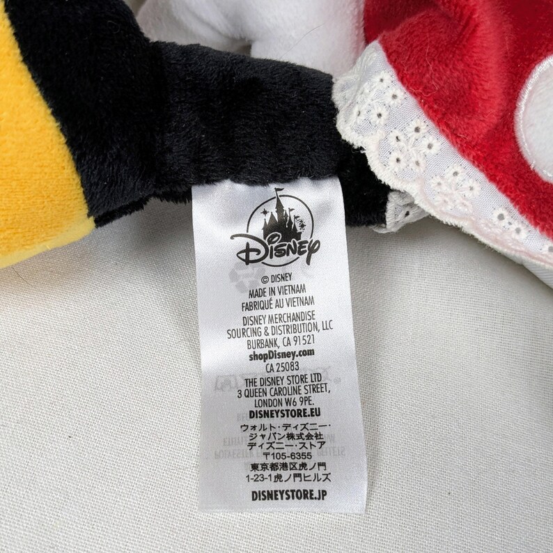 May include: A Disney product tag with the Disney logo, Made in Vietnam text, and various address details. The tag is attached to a plush toy with black, yellow, and red fabric.