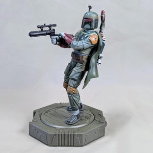 Pu&ograve; includere: Statuetta dettagliata di Boba Fett, un personaggio di Star Wars, in posa dinamica. La figura indossa un'armatura usurata con un mantello verde, che tiene un fucile blaster. Si trova su una base esagonale, mostrando dettagli intricati e un'estetica consumata dalla battaglia.