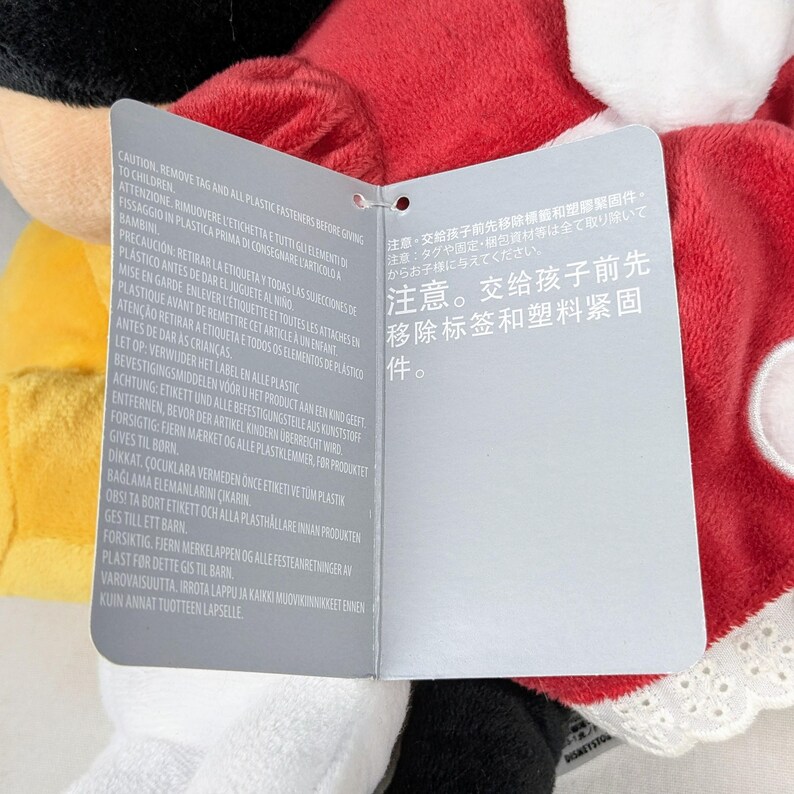 May include: A plush Minnie Mouse doll wearing a red dress with white polka dots and a white collar. A tag with multilingual safety instructions is attached to the doll.