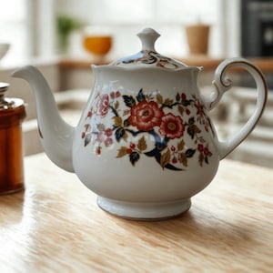 May include: White ceramic teapot with a floral pattern in red, blue, and gold. The teapot has a curved spout, a rounded body, and a lid with a small knob. The handle is curved and has gold trim. A classic tea accessory.