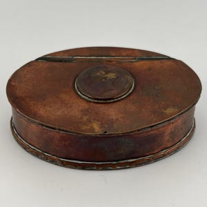 May include: An oval-shaped copper trinket box with a raised circular design on the lid. The box has a white and copper trim around the edge.